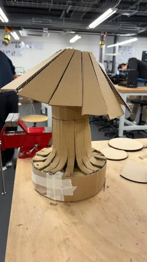 Cardboard lo-fi lamp model with a pleated shade on a workshop bench